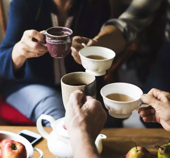 Four different hands holding four different cups of tea with fruits and kettle on the tea table
