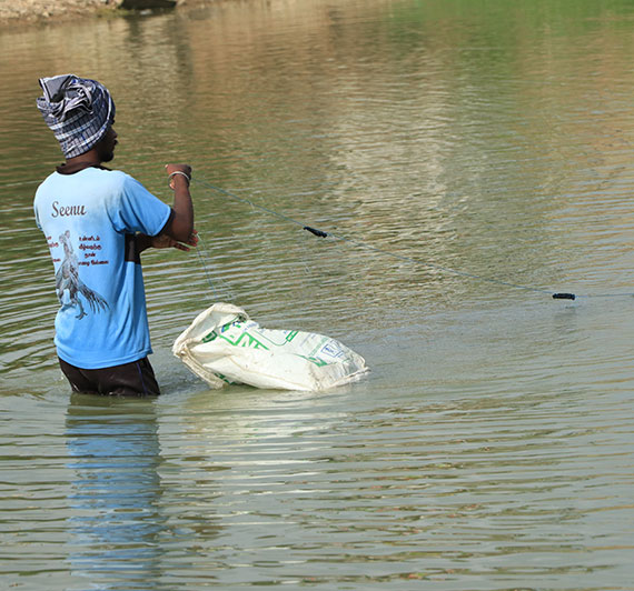 A man standing in the middle of a lake throwing a fishing net with a plastic sack floating nearby