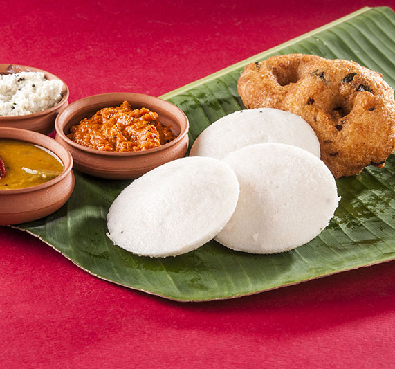 An inviting breakfast of soft idlis and vadas served on a banana leaf with sambar, coconut chutney, and kaara chutney on three different mud bowls